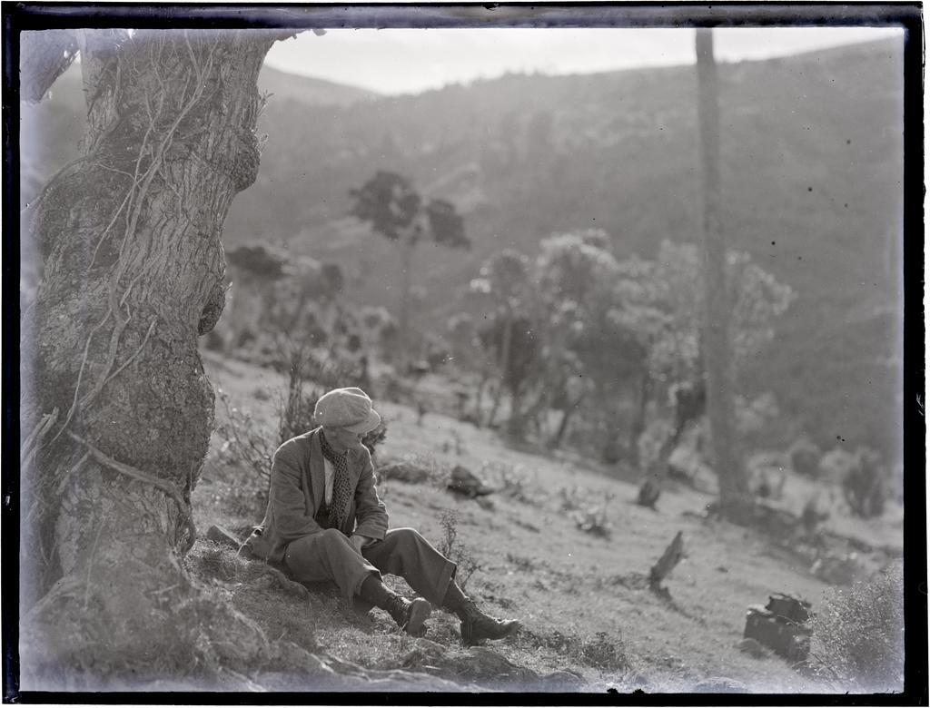 Young man wearing a cloth cap and scarf, sitting on a hillside under a tree