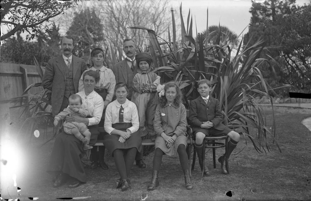 Group in front of flax bush, Vic McGeorge on Miss Gillies' knee