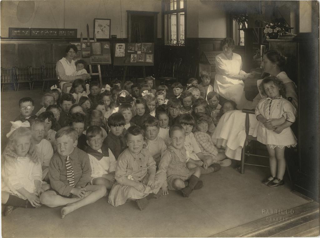 Children (mostly seated) with three women in uniform