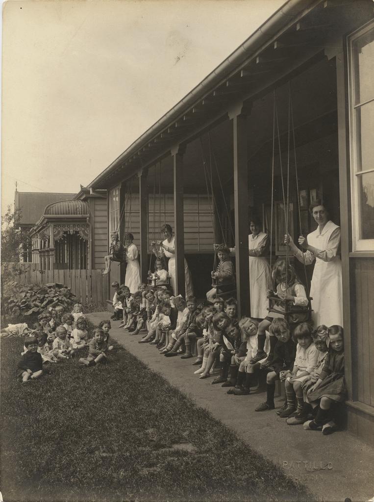 Children on lawn and kindergarten verandah, with four women in uniform