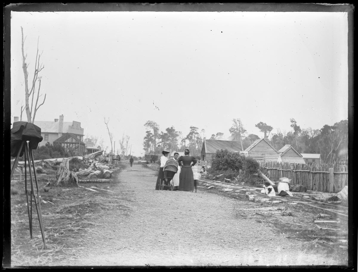 Scene [at Colac Bay] showing a road, tramway line, buildings, and a group of women and children