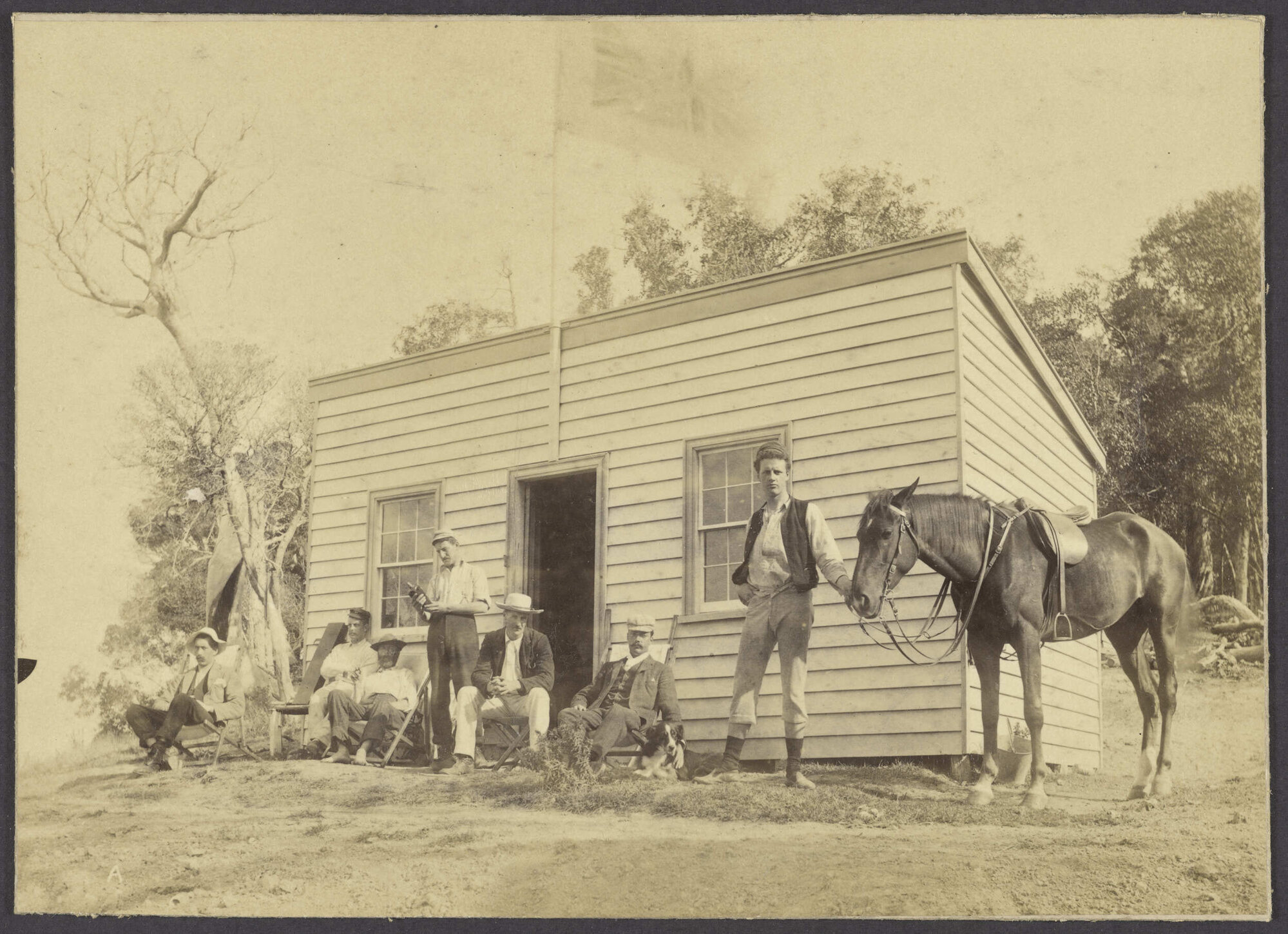 Group of men with horse and dog, in front of a small building