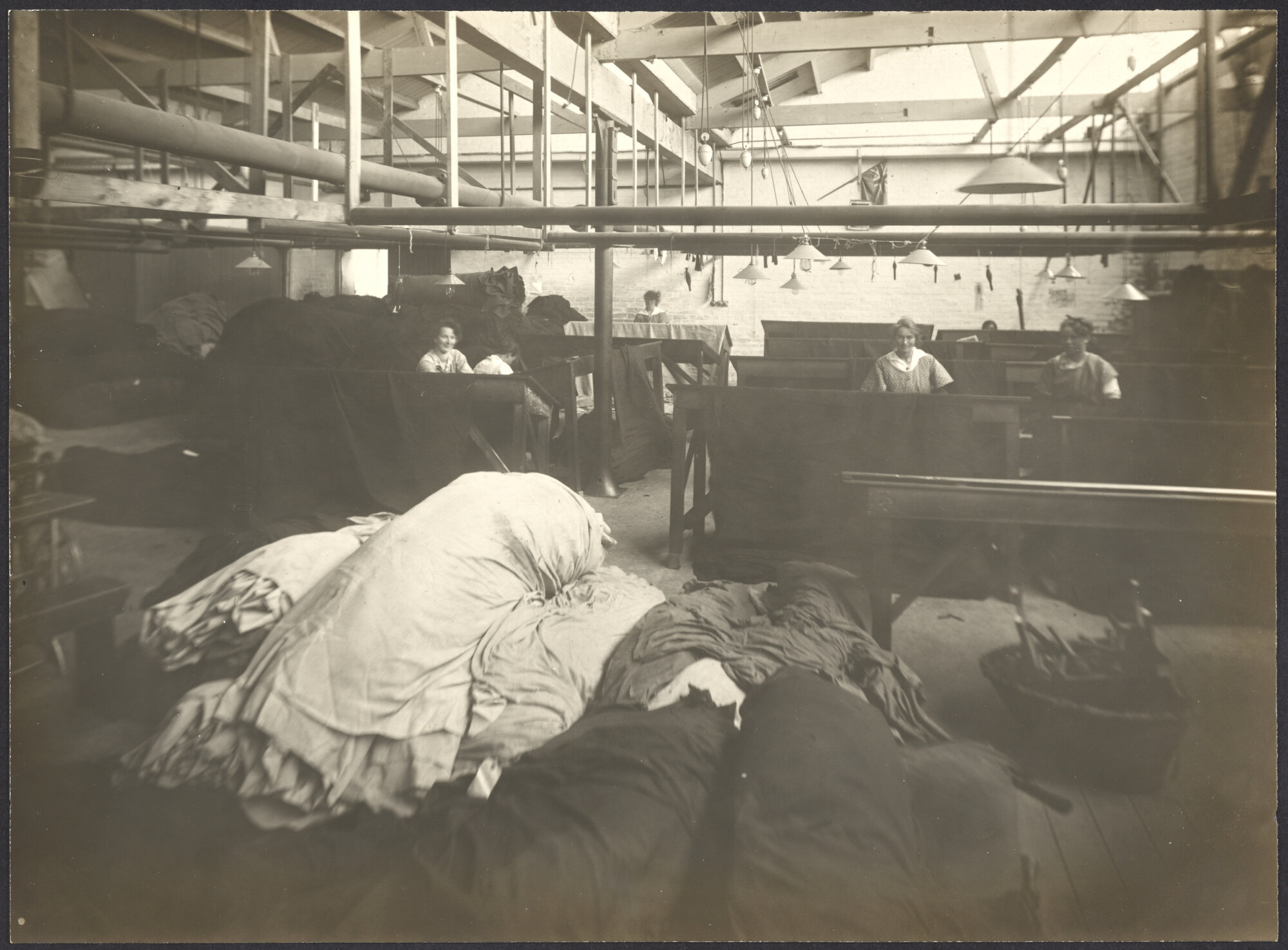 Women workers darning cloth at sloping tables, large pile of cloth in foreground