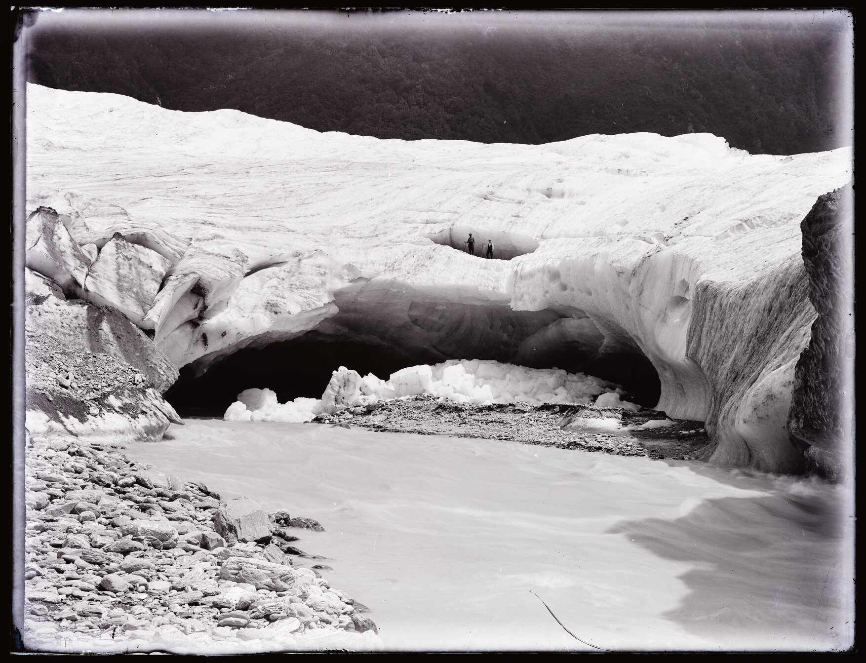 Two people standing on an arch of glacial ice next to a stream, Westland