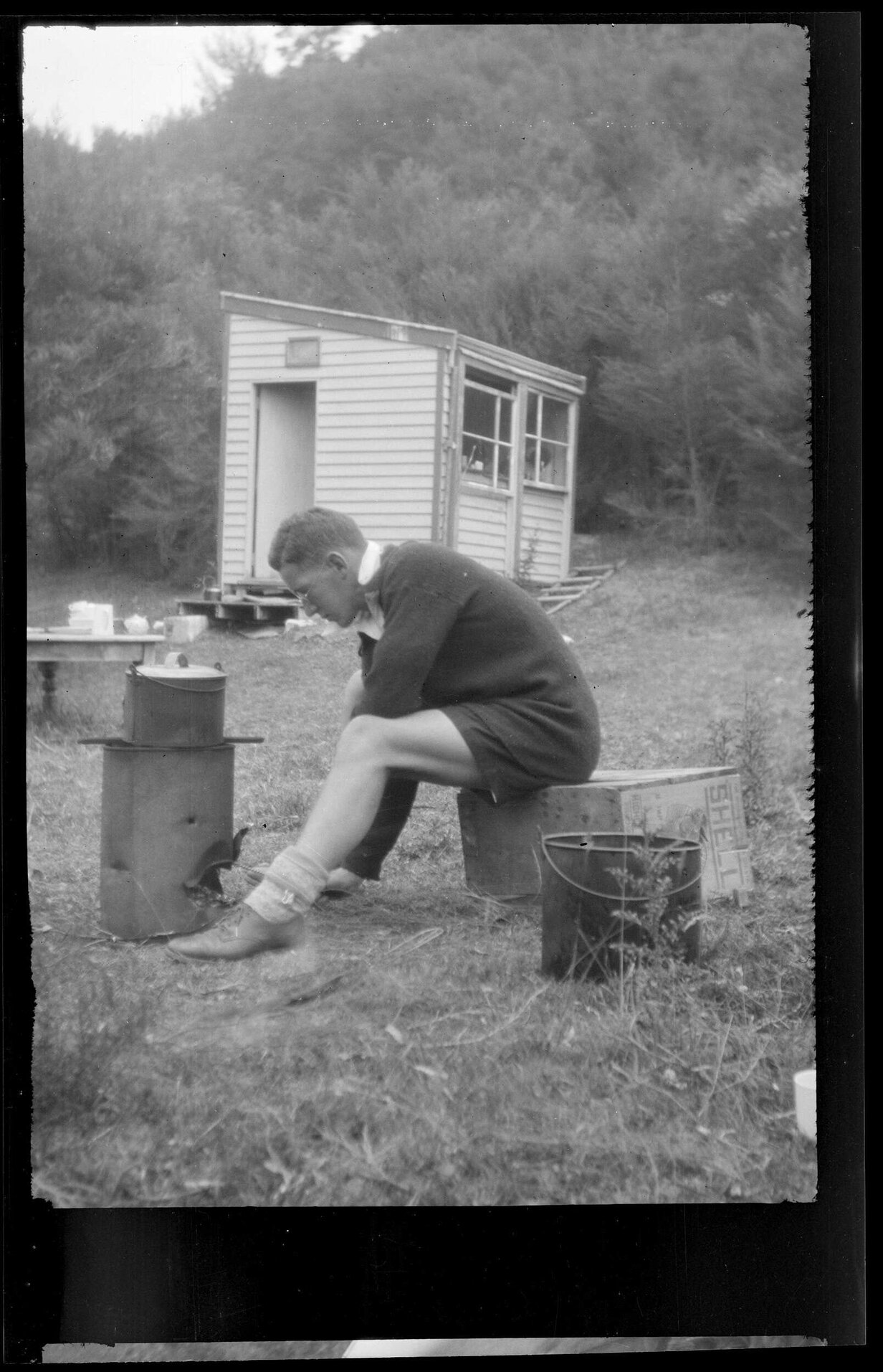 Man sitting on a crate at a camp site, with hut in background