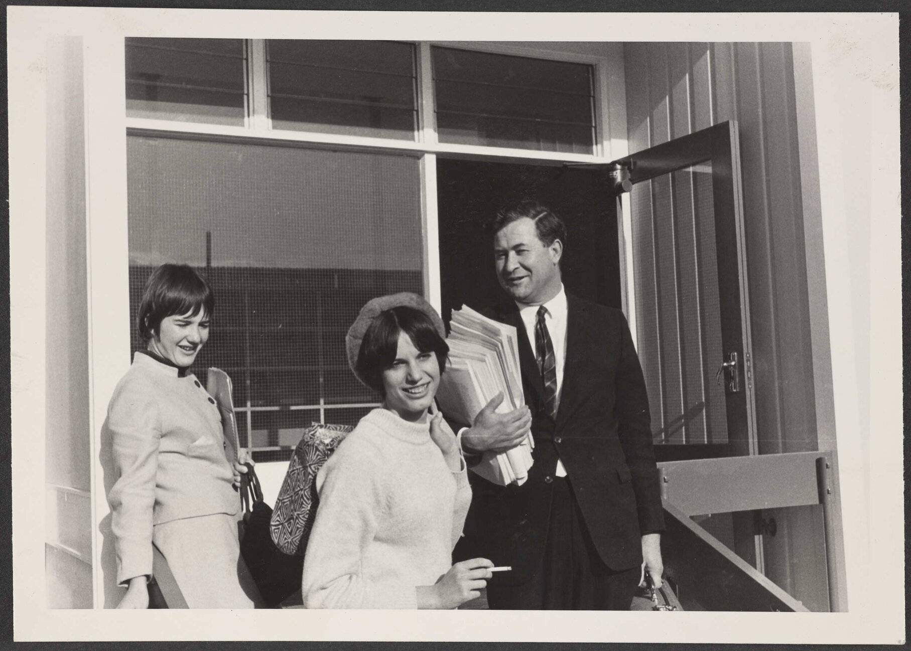Three people with bags and papers, standing on steps in front of doorway