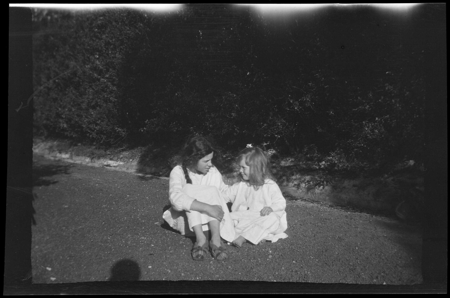 Two girls wearing smocks, sitting in front of a hedge