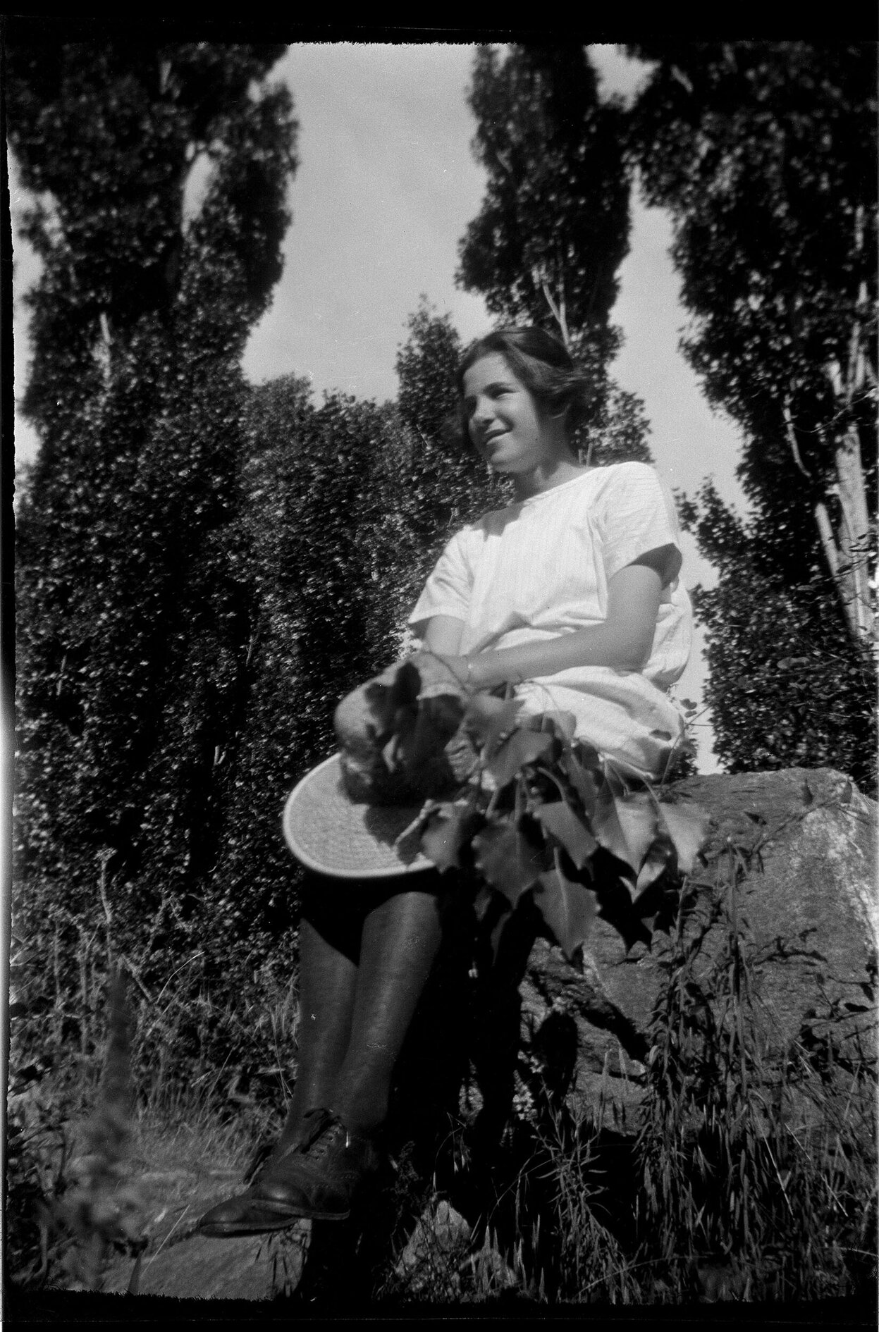 Lesley Brasch [?] in summer clothing, sitting on a rock and holding a hat