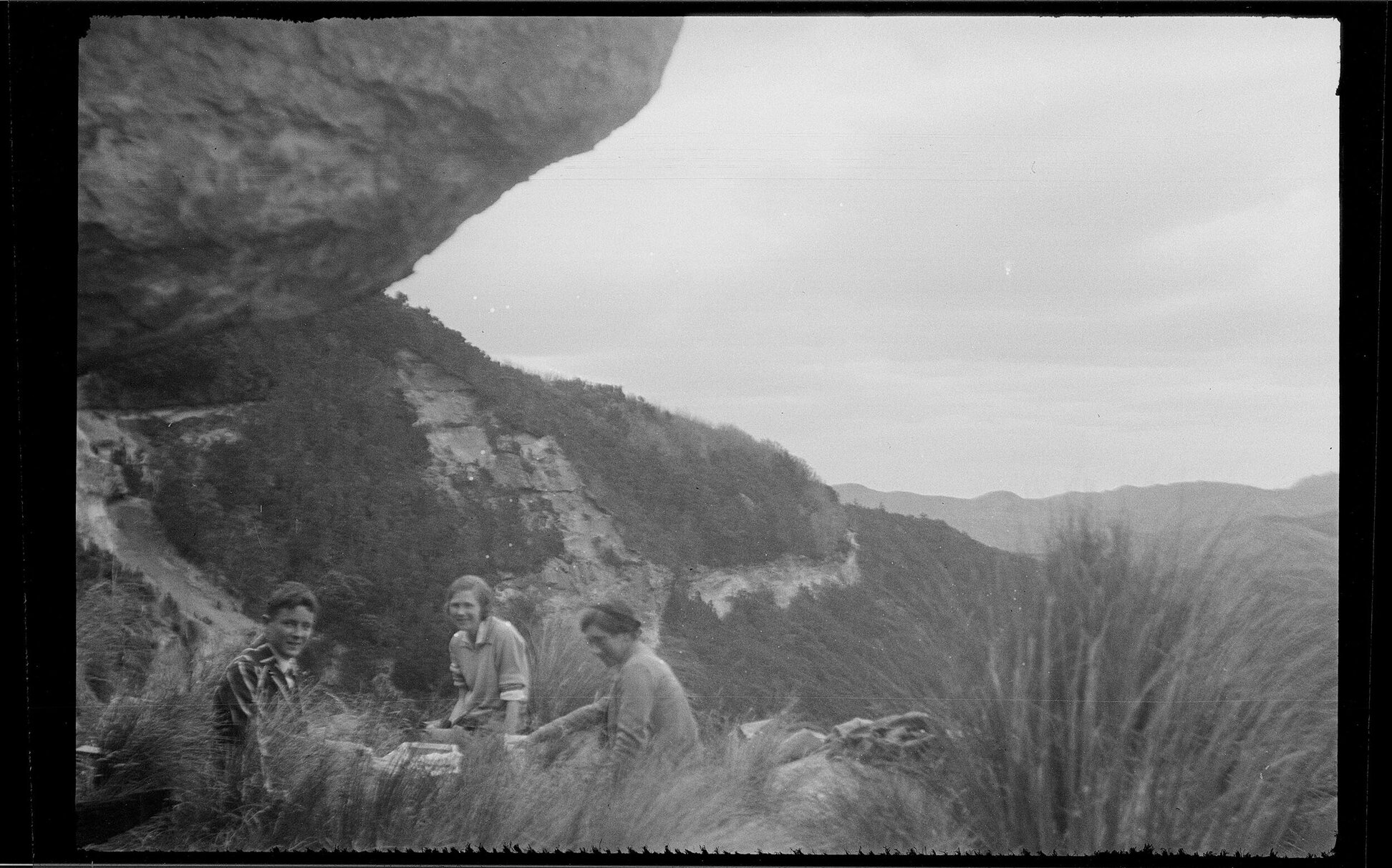 Three people sitting beneath a large rock in rugged terrain