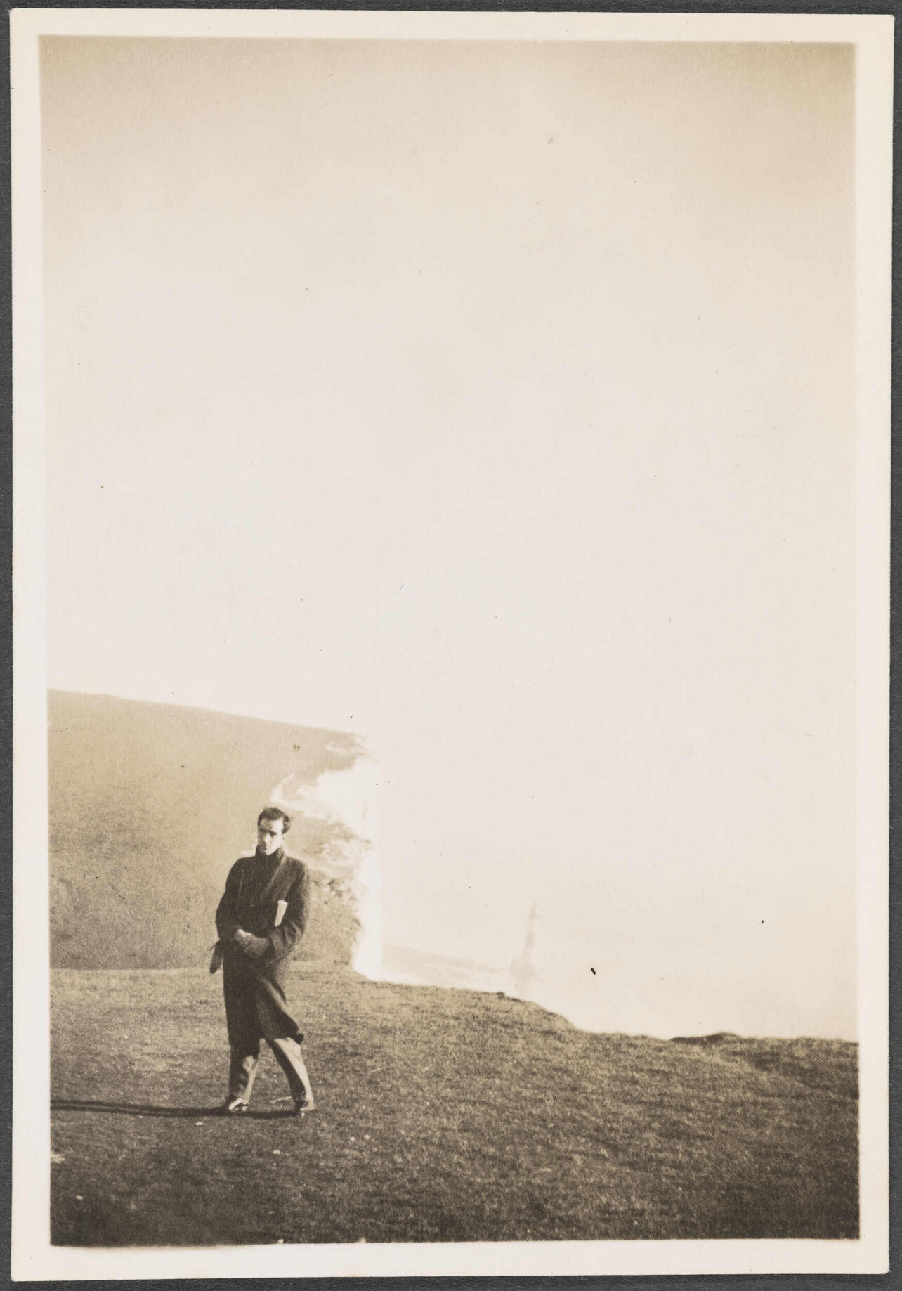 Charles Brasch standing in front of cliffs with a lighthouse in the distance