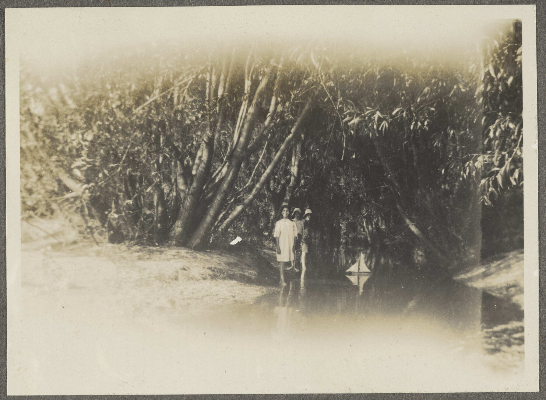Three children standing in a creek with toy sailing boat