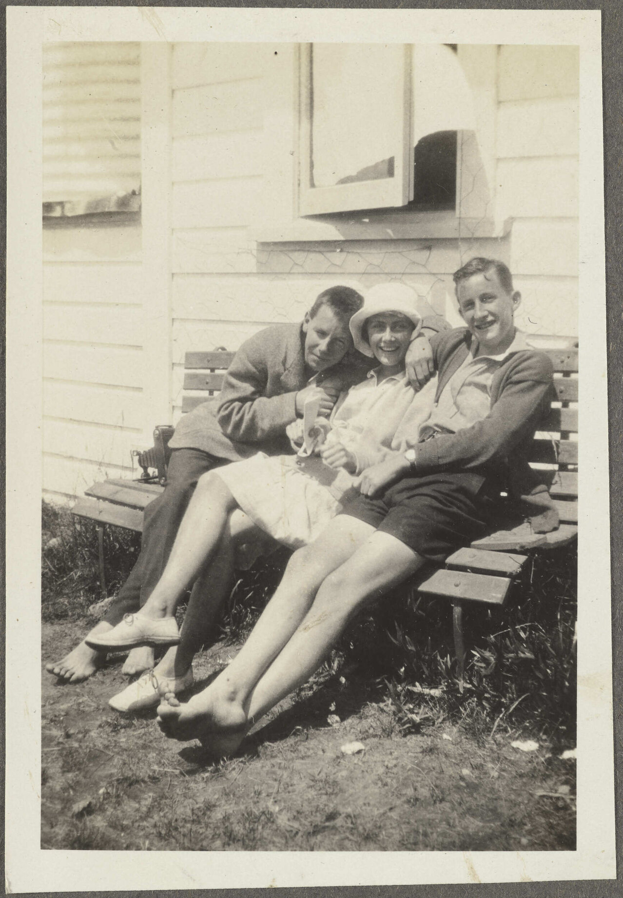 Two unidentified men and one woman sitting on bench outside wooden house
