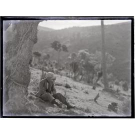 Young man wearing a cloth cap and scarf, sitting on a hillside under a tree