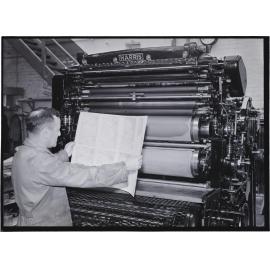 A man holding uncut pages [of a 'Shell Road Maps New Zealand book'] in front of a Harris printing press, Coulls Somerville Wilkie, Crawford Street, Dunedin