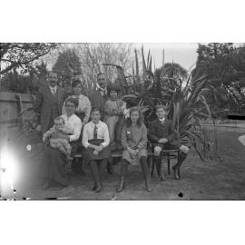 Group in front of flax bush, Vic McGeorge on Miss Gillies' knee