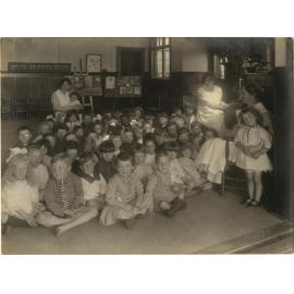 Children (mostly seated) with three women in uniform