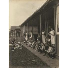 Children on lawn and kindergarten verandah, with four women in uniform
