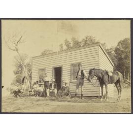 Group of men with horse and dog, in front of a small building