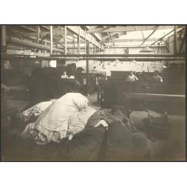 Women workers darning cloth at sloping tables, large pile of cloth in foreground
