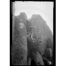 Charles Brasch sitting between large rocks