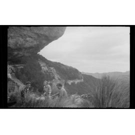 Three people sitting beneath a large rock in rugged terrain
