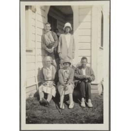 Four unidentified women and two men sitting outside wooden house