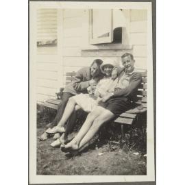 Two unidentified men and one woman sitting on bench outside wooden house