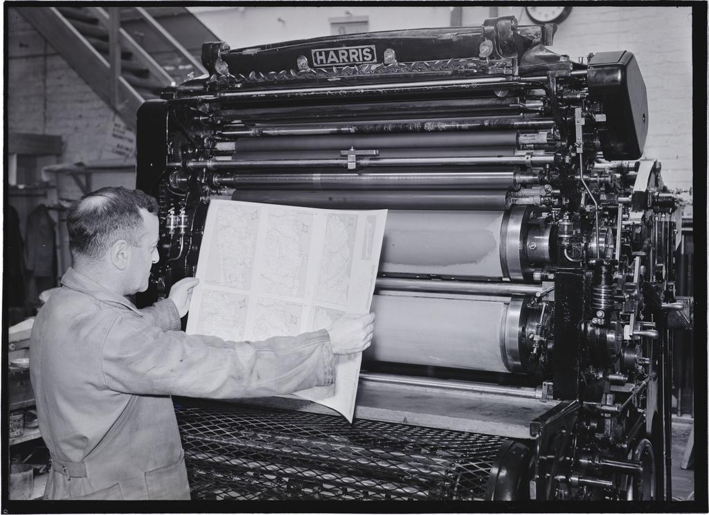 A man holding uncut pages [of a 'Shell Road Maps New Zealand book'] in front of a Harris printing press, Coulls Somerville Wilkie, Crawford Street, Dunedin